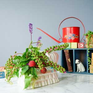 May include: A white ceramic planter shaped like a bed with a floral pattern, filled with green leaves, purple flowers, and red berries. A red watering can with floral designs, a wooden shelf with colorful blocks, and a small white dog figurine are in the background.