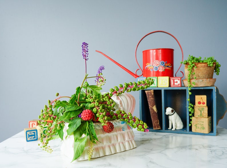 May include: A white ceramic planter shaped like a bed with a floral pattern, filled with green leaves, purple flowers, and red berries. A red watering can with floral designs, a wooden shelf with colorful blocks, and a small white dog figurine are in the background.