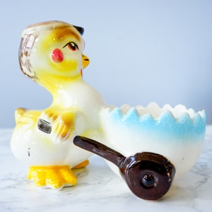 May include: A ceramic chick figurine pushing a blue and white egg-shaped cart. The chick is yellow with brown and black markings and red cheeks. The cart has a brown wheel.