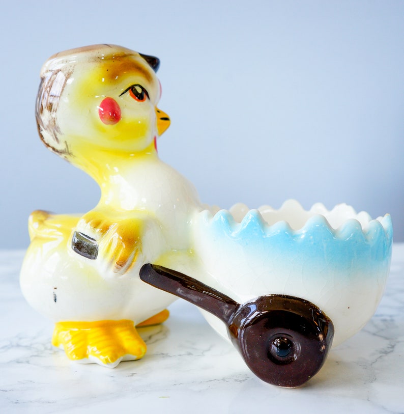 May include: A ceramic chick figurine pushing a blue and white egg-shaped cart. The chick is yellow with brown and black markings and red cheeks. The cart has a brown wheel.