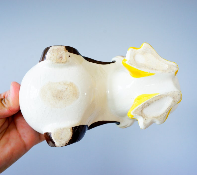 May include: White ceramic planter shaped like a dog with brown and yellow accents. The planter is lying on its side, showing the bottom of the planter.