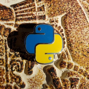 Op de afbeelding: Een gele en blauwe emaille pin in de vorm van het logo van de programmeertaal Python.