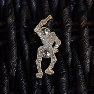 May include: Silver-toned metal pin featuring a skeleton figure holding a bat, with a detailed design and two silver-toned metal fasteners on the back.