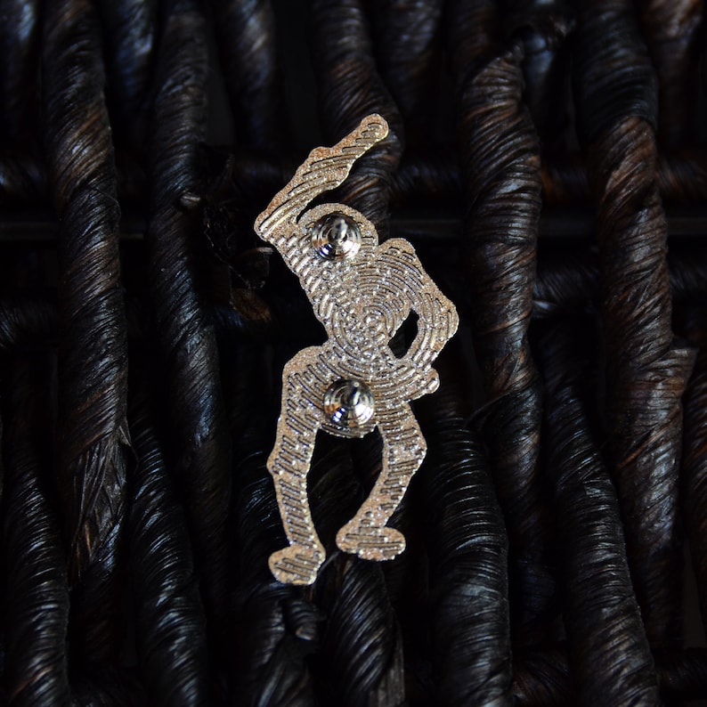 May include: Silver-toned metal pin featuring a skeleton figure holding a bat, with a detailed design and two silver-toned metal fasteners on the back.