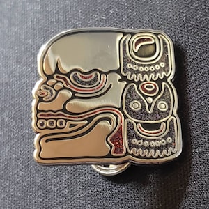May include: Silver metal pin featuring a detailed design of a skull and a stylised face with red and black accents. The pin has a shiny finish.