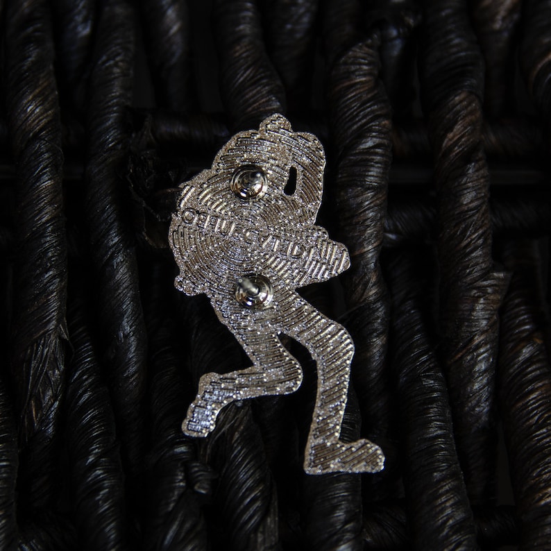 May include: A silver-toned metal pin depicting a cartoon-like figure in a running pose. The pin has a detailed design and a textured surface. The back of the pin features two metal posts for attaching it to clothing or a bag.