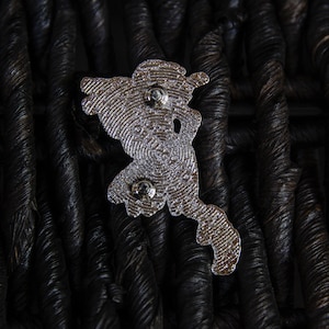 May include: Silver metal pin featuring a detailed design of a cartoon character. The pin has two metal posts on the back for attaching to clothing or a bag.