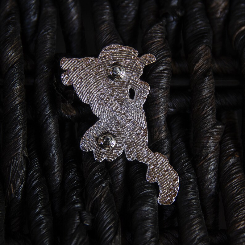 May include: Silver metal pin featuring a detailed design of a cartoon character. The pin has two metal posts on the back for attaching to clothing or a bag.