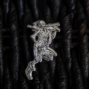 May include: Silver-toned metal pin depicting a detailed skeletal figure holding a weapon, set against a dark woven background.