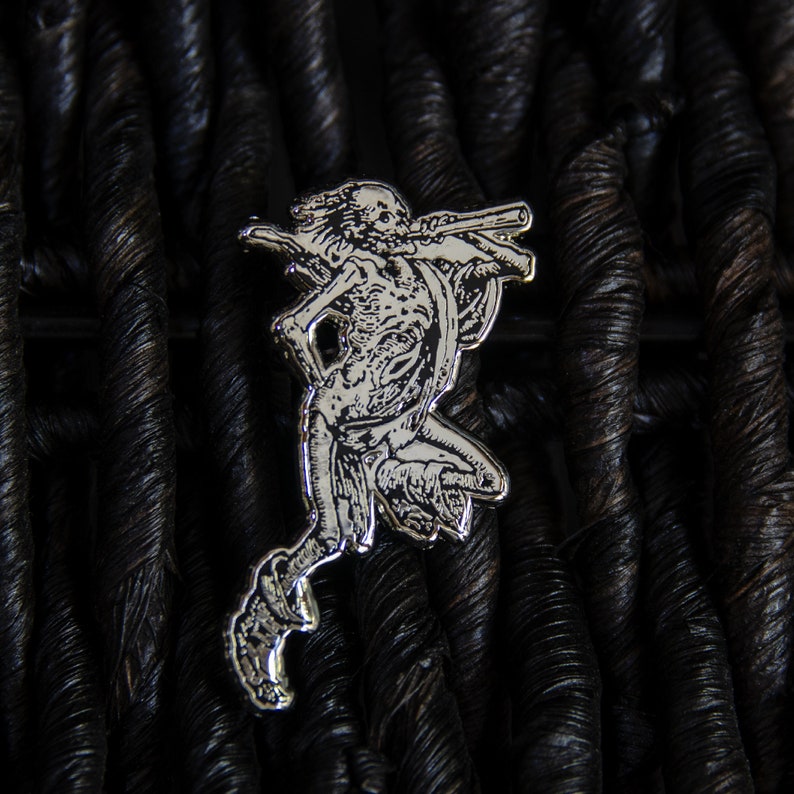 May include: Silver-toned metal pin depicting a detailed skeletal figure holding a weapon, set against a dark woven background.