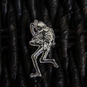 May include: Silver-toned metal pin depicting a skeleton playing a guitar. The skeleton is in a dynamic pose, with its head tilted back and its arms raised. The pin is set against a dark, textured background.