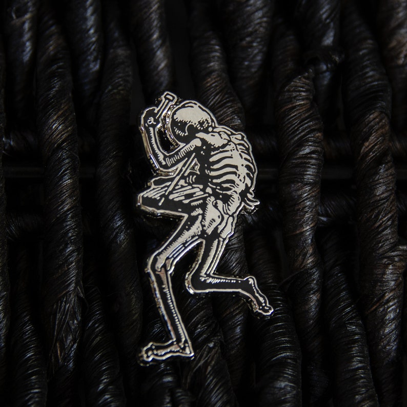 May include: Silver-toned metal pin depicting a skeleton playing a guitar. The skeleton is in a dynamic pose, with its head tilted back and its arms raised. The pin is set against a dark, textured background.