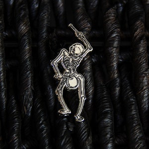 May include: A silver skeleton pin holding a drumstick, set against a black woven background.