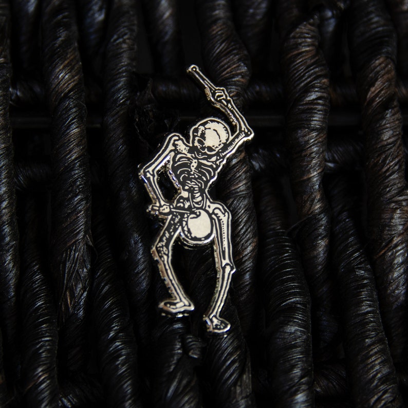 May include: A silver skeleton pin holding a drumstick, set against a black woven background.
