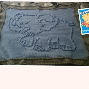 May include: A blue crocheted baby blanket with a raised elephant design. The blanket has a green border. The text "Grr-ific! Graphs & Gifts" is visible in the upper right corner of the image.