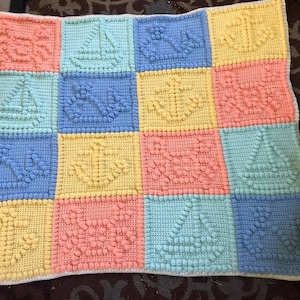 Könnte beinhalten: Eine gehäkelte Babydecke mit einem bunten Patchwork-Muster. Jedes Quadrat zeigt ein anderes strukturiertes Muster, darunter Segelboote, Herzen und Blumen. Die Decke ist aus weichen Pastellfarben gefertigt, darunter Gelb, Blau, Rosa und Grün.