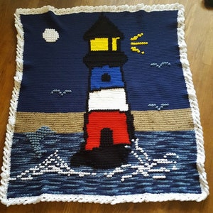 Op de afbeelding: Een gehaakt deken met een vuurtoren met een geel licht, rode, witte en blauwe strepen en een zwarte basis. De vuurtoren staat op een zandstrand met blauwe golven en wit schuim. De achtergrond is een donkerblauwe lucht met een witte maan en een gele zon.