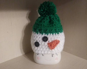 May include: A white crocheted snowman with a green hat and a red nose. The snowman has black button eyes and a white pom-pom on top of the hat.