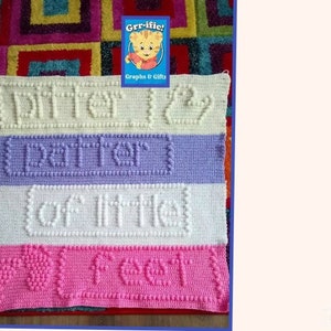 以下が含まれることがあります： 異なる色の4本のストライプが施された、かぎ針編みのベビーブランケット。一番上のストライプはクリーム色で、「Ditty 129」という文字が浮き彫りになっています。2番目のストライプはラベンダー色で、「partner」という文字が浮き彫りになっています。3番目のストライプはクリーム色で、「of little」という文字が浮き彫りになっています。一番下のストライプはピンク色で、「feet」という文字が浮き彫りになっています。このブランケットは、浮き彫りの効果を生み出すテクスチャのあるステッチで編まれています。