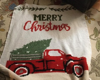 May include: A crocheted Christmas blanket featuring a red truck carrying a green Christmas tree. The words "MERRY Christmas" are displayed in red and green. The blanket is primarily off-white with a textured appearance.