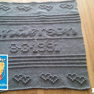 May include: A gray knitted baby blanket with the words "Grr-ific!" and "Baby" in raised letters. The blanket features heart-shaped designs and horizontal stripes. A small graphic of a cartoon tiger is in the corner.