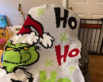 May include: A crocheted blanket featuring the Grinch from the movie "How the Grinch Stole Christmas". The blanket is gray with green, red, white, and black accents. The words "Ho Ho Ho" are written in large letters on the blanket.