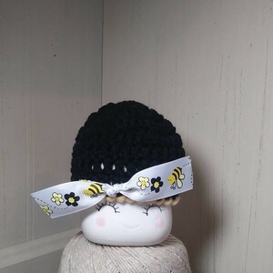 May include: Black crocheted baby hat with a white ribbon featuring yellow flowers and bees.