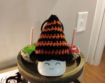 May include: A two-tiered wooden serving tray with a white ceramic mug with a smiling face and a black and orange crocheted witch hat on top. The tray is decorated with green and red candy apples with striped straws.