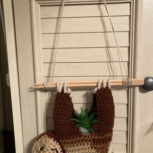 May include: A crocheted sloth hanging planter with a small potted plant inside. The sloth is brown and beige and has a small face with eyes. The planter is hanging from a wooden dowel rod with twine.