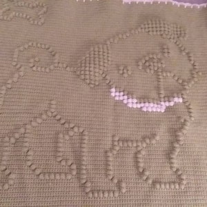 Op de afbeelding: Een groene gehaakte deken met een wit gehaakt ontwerp van een hond. De deken is gemaakt met een textuur-steek, waardoor het een pluche uitstraling heeft.