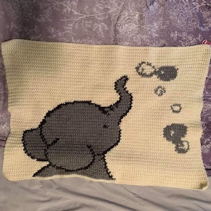 Puede incluir: Una manta de bebé de ganchillo con un elefante gris que sopla burbujas. La manta es de color crema con un borde gris.