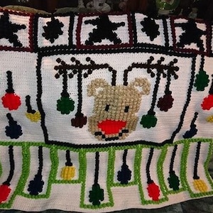 May include: A white crocheted blanket with a reindeer design and a border of green, black, and red ornaments. The reindeer has a brown nose and red antlers.