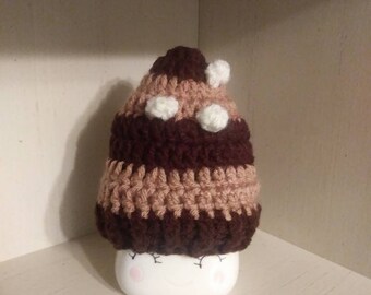 May include: A handmade crochet hat in shades of brown and tan, topped with white pom-poms, sits atop a white ceramic container with a smiling face. The hat features a striped pattern and is likely a decorative item.