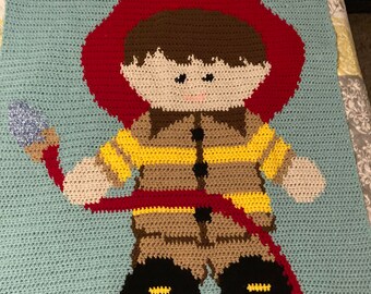 May include: A crocheted baby blanket featuring a cartoon firefighter with a red hat, yellow jacket, and brown boots. The blanket is light blue with a white border.