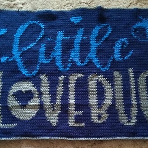 May include: A blue crocheted blanket with the words "Little Loved" in white and blue yarn. A small blue and white crocheted design is in the top right corner.