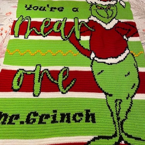 May include: A rectangular crochet blanket featuring horizontal stripes in red, white, and lime green. The blanket displays a graphic of a green character in a red Santa suit and the text "You're a mean one Mr. Grinch" in black and green lettering.