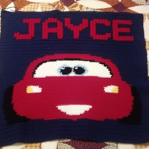 Puede incluir: Manta de ganchillo hecha a mano con un diseño de coche de carreras rojo sobre un fondo azul marino. El nombre "JAYCE" se muestra en letras rojas encima del coche. La manta es probablemente para un niño.