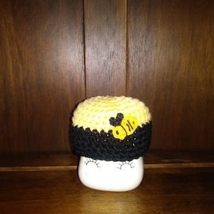 May include: A yellow and black crocheted baby hat with a bee design. The hat is sitting on a white mug with black eyelashes.