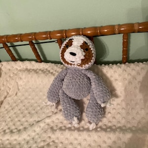 May include: A handmade crochet sloth toy in shades of gray, brown, and white. The sloth has a round head with a brown face and white accents. Its body and limbs are a soft gray, with white details on the feet and hands. The toy is resting on a white, textured blanket.