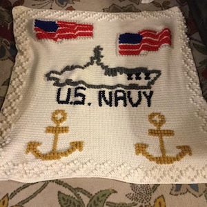 May include: White crocheted blanket with a navy blue submarine, two gold anchors, and two American flags. The blanket has a white crocheted border with small white pom-poms.