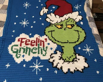 May include: A blue crocheted blanket with a green Grinch character wearing a red Santa hat. The blanket has white snowflakes and the text "Feelin' Grinchy" in green and white.