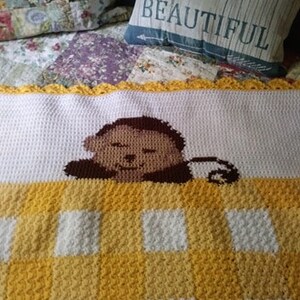 Könnte beinhalten: Eine weiße und gelbe gehäkelte Babydecke mit einem braunen Affengesicht in der Mitte. Die Decke hat einen gelben gehäkelten Rand.