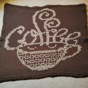 May include: A brown crocheted blanket with a beige crocheted design of a coffee cup with steam rising from it. The design says "Coffee" in stylized letters.