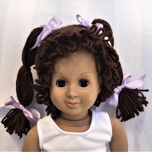 May include: A brown yarn doll wig with two pigtails tied with lavender bows. The wig is styled in loose curls.