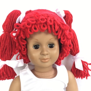 May include: A red yarn wig with white bows for a doll. The wig has curly red yarn hair with two pigtails tied with white bows.