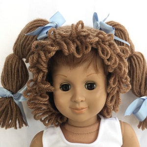 May include: A doll with brown yarn hair styled in pigtails with blue ribbon bows. The doll is wearing a white tank top.