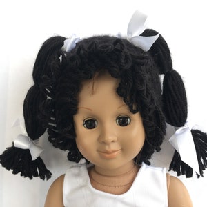 May include: A doll with black curly yarn hair styled in pigtails with white bows. The doll is wearing a white sleeveless shirt.