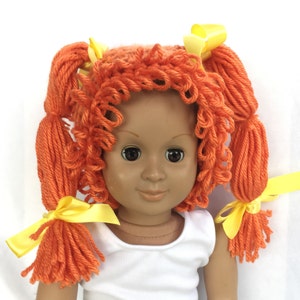 May include: A doll with orange yarn hair styled in pigtails with yellow bows. The doll has brown eyes and is wearing a white tank top.