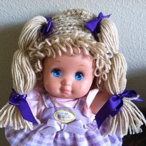 May include: A doll with blonde yarn hair styled in pigtails with purple bows. The doll is wearing a purple and white gingham dress with a pink undershirt and a purple bib with the words "Magic Nursery" on it.
