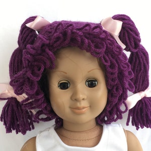 May include: Purple yarn doll wig with pink bows. The wig has two pigtails and is styled in loose curls.